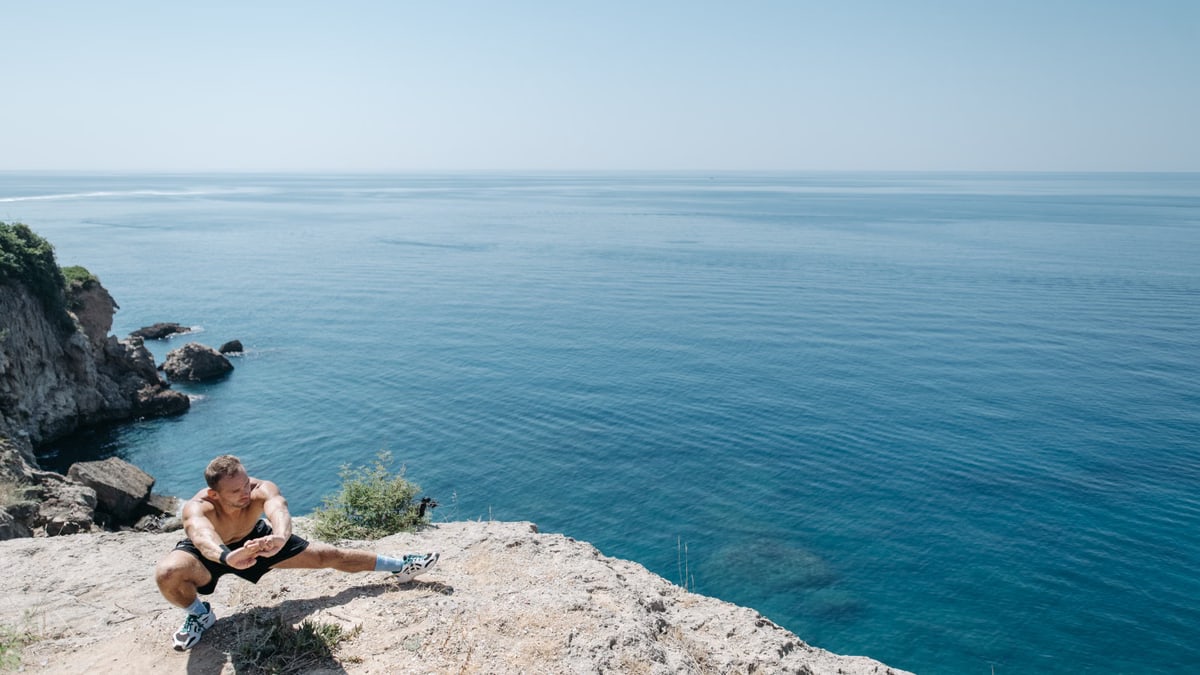 Shirtless man stretching on a cliff's edge by the ocean showcasing a healthy lifestyle and fitness.